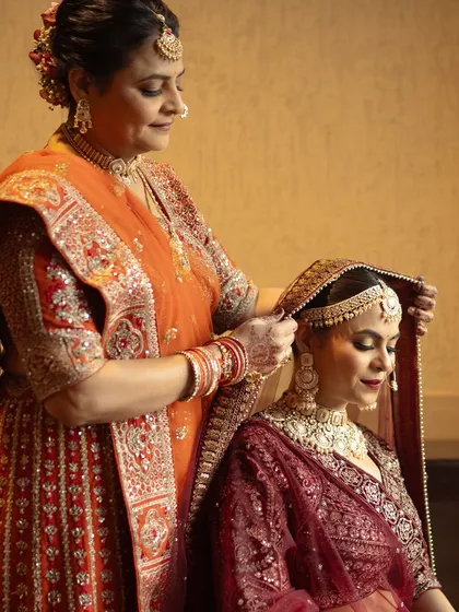 A touching moment between a mother and daughter. The bride, in a stunning deep red lehenga, gets her final touches, her makeup looking flawless and regal.