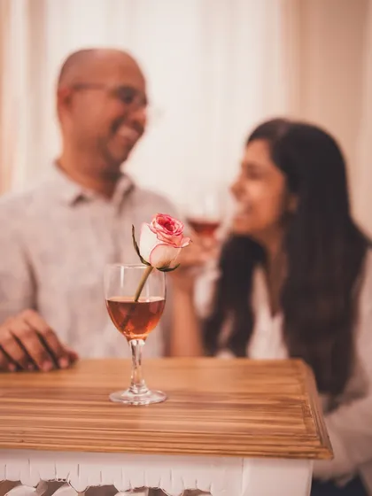 A creative shot focusing on a wine glass with a rose, with a happy couple blurred in the background. This tells a story of love and celebration.