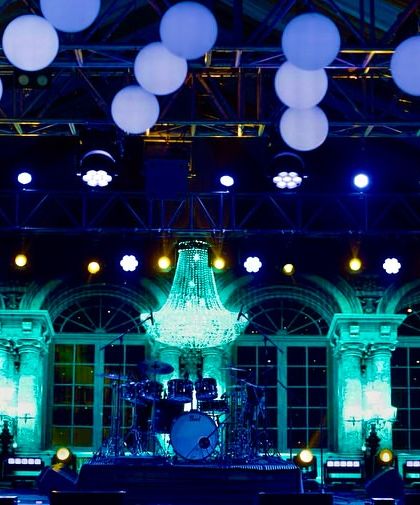 The stage setup for the band, with a stunning crystal chandelier and a backdrop of classic architectural facades lit in cool blue tones, blending classic elegance with a modern party vibe.