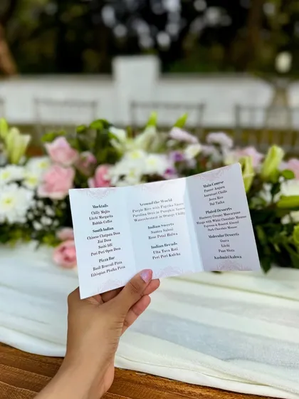 A hand holds up a beautifully designed, tri fold menu against a backdrop of fresh flowers. This highlights my service of creating custom menus that match the theme and elegance of the event.