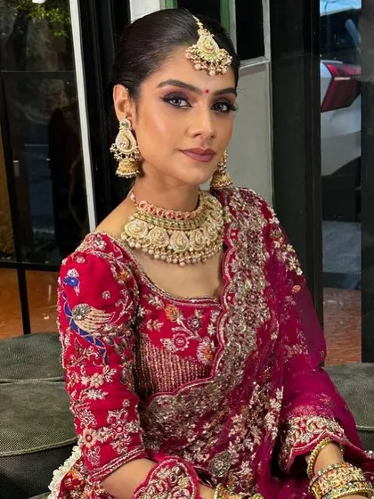A full view of this stunning bridal look. The makeup is balanced to complement the heavy embroidery of the lehenga and the intricate jewelry, with a sleek hairstyle to keep the focus on her face.