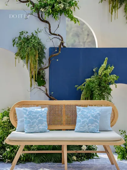 The main seating area for the Rishikesh Mehndi. A rattan bench with blue cushions is set against a modern, layered backdrop of blue and white arches, adorned with organic branches and greenery.