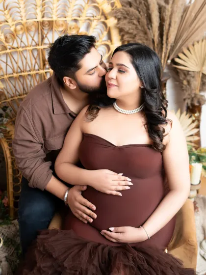 A tender moment captured in a bohemian-styled maternity shoot. The warm, earthy tones of the outfits and the natural texture of the wicker chair and pampas grass create a cozy, intimate atmosphere.