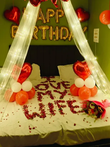 A sweet and personal birthday message, "I LOVE MY WIFE," written in rose petals. The bed is framed by a light canopy and heart-themed balloon bouquets.