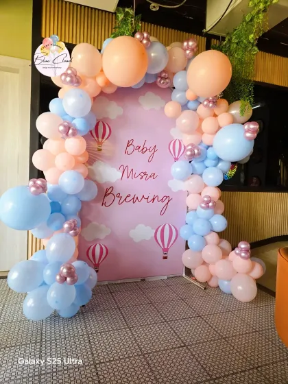 A hot air balloon themed baby shower with a 'Baby Misra Brewing' custom backdrop and a pink and blue balloon garland.