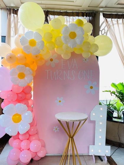 A cheerful daisy-themed first birthday party. The pink backdrop is framed by a pink, yellow, and white balloon arch with cute daisy flower balloon accents.