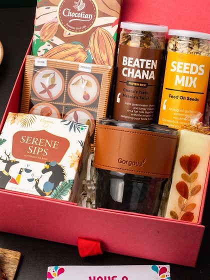 This gift box is designed for a cozy and comforting experience. It features a stylish glass tumbler with a leather-finish grip, healthy snacks like beaten chana and seeds mix, serene sips tea, and artisanal chocolates, perfect for a relaxing evening.