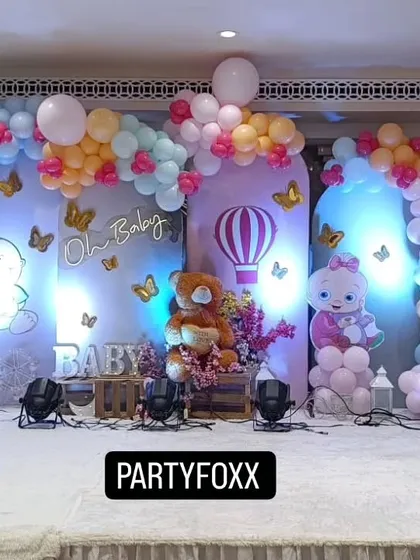 A cute baby shower theme with pastel balloon arches, character cutouts, a central teddy bear prop, and a hot air balloon detail.