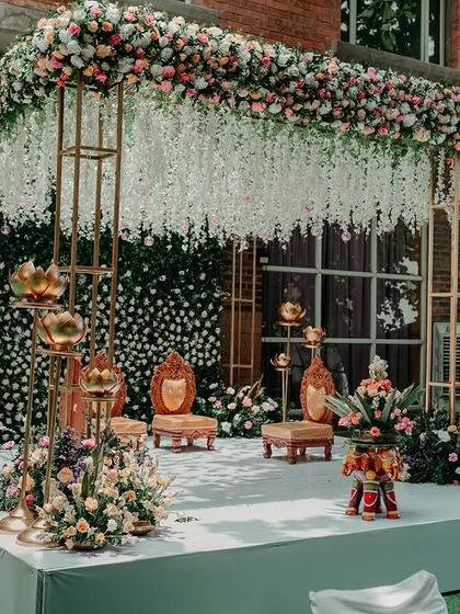 A modern mandap setup for a wedding expo, showcasing a gold frame, a wall of white flowers, and hanging floral installations to demonstrate our design capabilities.