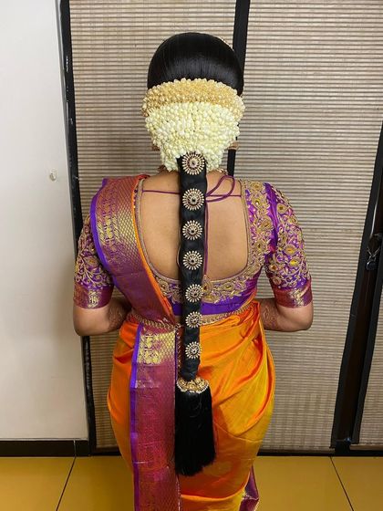 A classic bridal braid with circular gold ornaments and a full jasmine veni. This hairstyle is a perfect match for the bride's vibrant orange and purple Kanjivaram saree.