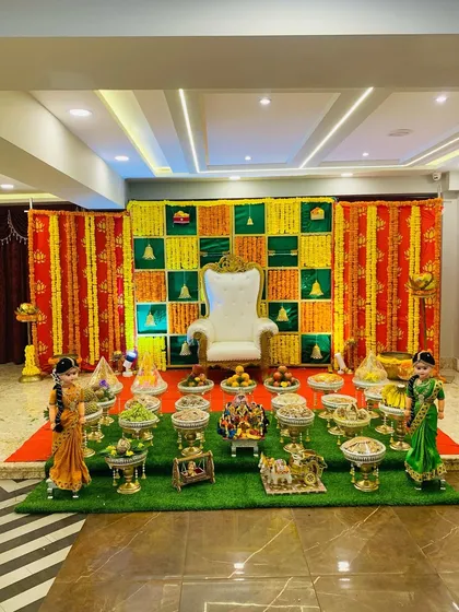 A full view of a traditional Seemantha setup with a green lawn, offering trays, and a colorful checkerboard backdrop. The hanging garlands and traditional dolls complete this festive scene.