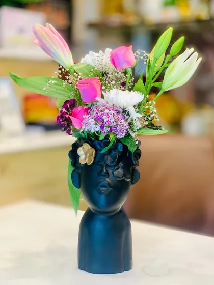 A modern and artistic floral arrangement in a unique face-shaped vase. This piece combines fresh lilies and roses, but I can also create a similar long-lasting version with artificial flowers.