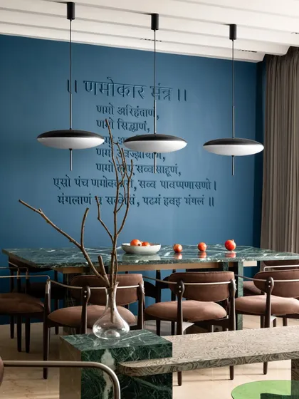 The dining area, where a green Indian marble table is set against a deep blue wall featuring a Jain mantra. The mantra's letters are finished in the same tone as the wall, creating an understated yet powerful spiritual element in this modern space.