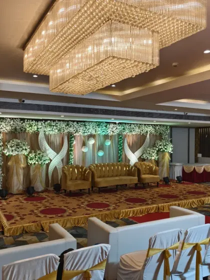 A luxurious stage design with golden sofas and a backdrop combining white floral arrangements with elegant drapery. The grand crystal chandelier adds to the opulent feel of the venue.