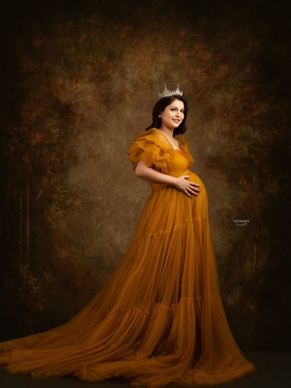 A regal portrait of a mom-to-be wearing a crown with her flowing mustard tulle gown. The classic painted backdrop adds a touch of royal sophistication to the image.