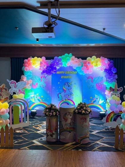 A full stage setup for a unicorn-themed birthday party. The backdrop features unicorns against a starry sky, illuminated by spotlights, with rainbow balloon columns and themed pedestals.