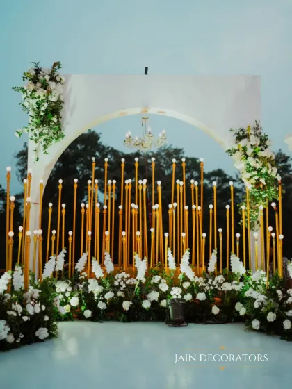 A modern and artistic backdrop. A white arch is set before a wall of golden tubes with bulb lights, framed by lush white florals, creating a stunning contrast of textures and materials.