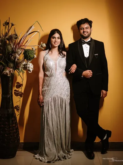 A classic, full-length portrait of the couple in their glamorous engagement outfits, showcasing their style against a warm, contemporary backdrop.