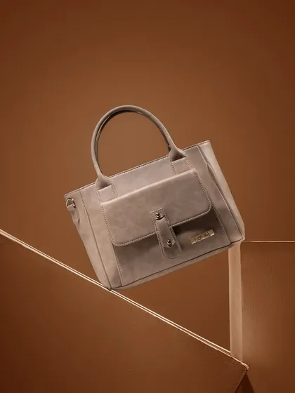 A monochromatic shot of a grey handbag against a background of geometric, cardboard-like shapes. The single-color palette is sophisticated and allows the bag's texture and form to take center stage.