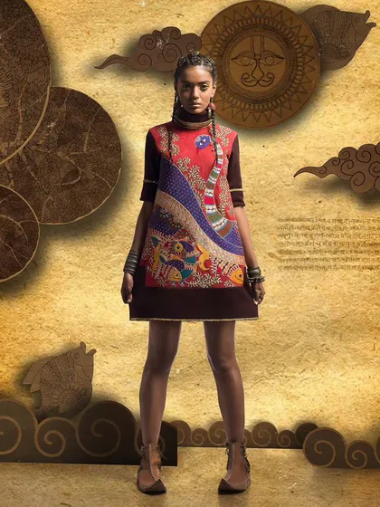 Full-length shot of a model in a short, art-inspired dress. The direct, centered pose against a textured, illustrative background makes the garment's unique peacock motif the central focus.
