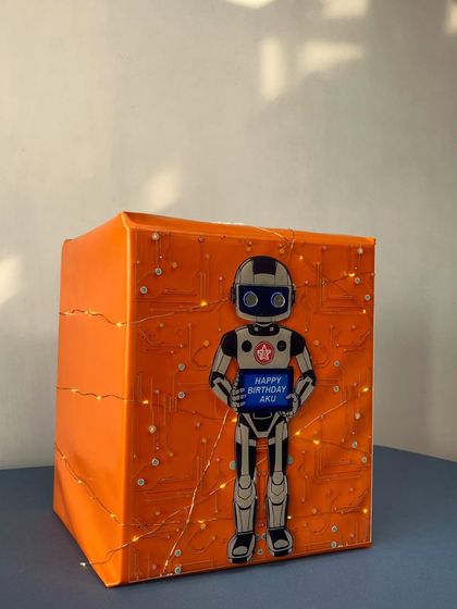 A futuristic, robot-themed gift box for a tech enthusiast. The bright orange wrap is detailed with a circuit board pattern and fairy lights, with a friendly robot holding a 'Happy Birthday' sign.