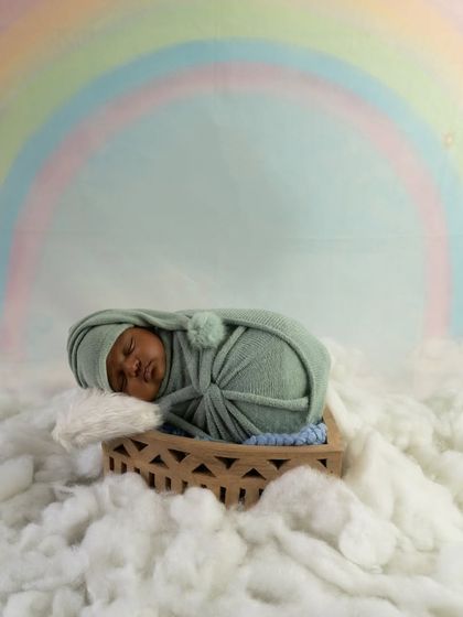 A newborn sleeps peacefully in a basket surrounded by soft, cloud-like cotton with a pastel rainbow in the background.