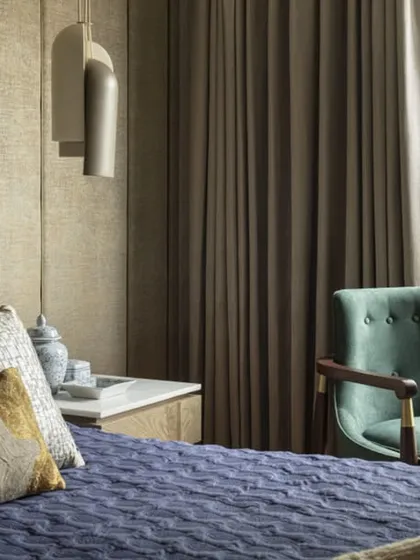A serene bedroom at the Jor Bagh Residence with a view of the lush green outdoors. The design combines a textured upholstered headboard, a deep blue quilted bedspread, and a comfortable armchair to create a peaceful escape.