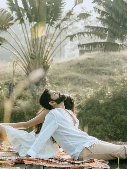 A relaxed, full-body shot of the couple on a picnic by the water, with large traveler's palms in the background. It has a very serene and tropical feel.