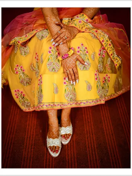 A top-down detail shot focusing on the bride's embroidered yellow lehenga, her henna-adorned hands, and her embellished footwear. I believe in capturing the beauty of the entire bridal ensemble.