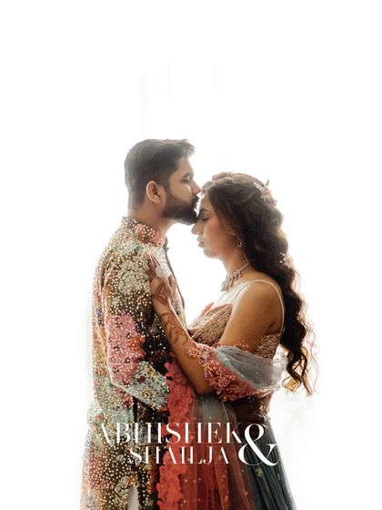 A tender forehead kiss against a bright, airy background. This high-key portrait highlights the couple's affection and the beautiful details of their Mehndi outfits.