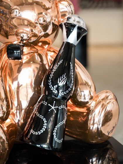 A close-up of the stylized bottle held by the "Let’s Party Teddy," featuring intricate designs on a black and silver surface.