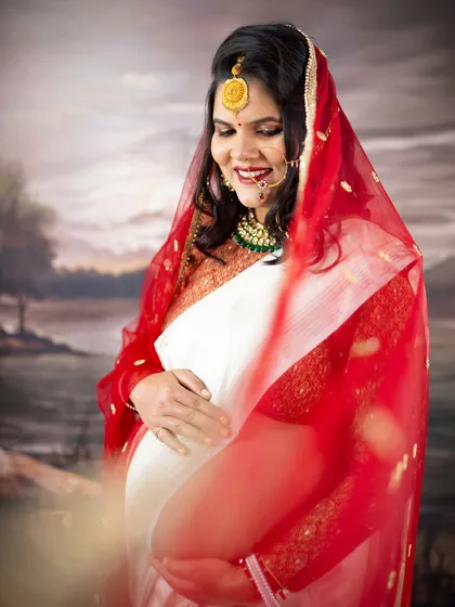 A beautiful portrait of a mom-to-be in a white and red saree, seated against a painted backdrop. The red veil adds a touch of traditional elegance.