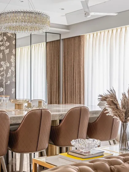 A close-up of the dining area, where the crystal chandelier casts a warm glow over the marble table. The composition highlights the luxurious textures of the quilted chairs and sheer curtains.