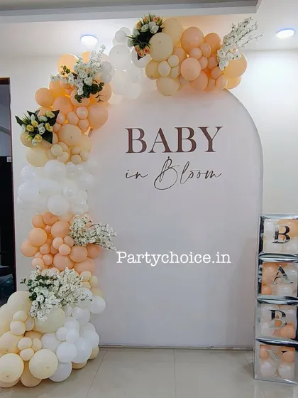A "Baby in Bloom" themed baby shower decor for an intimate home setting. This setup features a beautiful balloon arch in peach and white tones with floral accents.