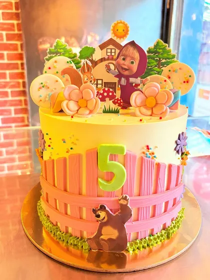 A charming Masha and the Bear cake, depicting a scene from the cartoon with a pink fence and forest elements.