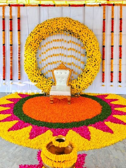 A vibrant and traditional puberty function setup, featuring a massive floral rangoli on the floor made from marigold petals. The backdrop includes a full floral ring and a classic white chair, creating a perfect photo opportunity.