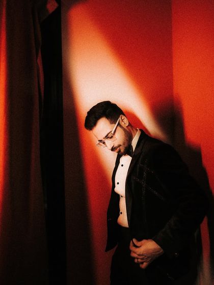 A moody, dramatic solo portrait of the groom under a spotlight. The deep red wall and strong shadows create a cinematic, almost theatrical feel.