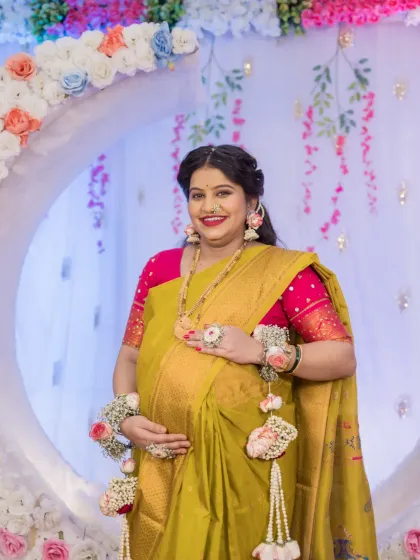 A beautiful solo maternity portrait of the mother-to-be, cradling her baby bump. She stands before a floral moon-gate, her traditional yellow saree and floral accessories perfectly complementing the festive baby shower decor.