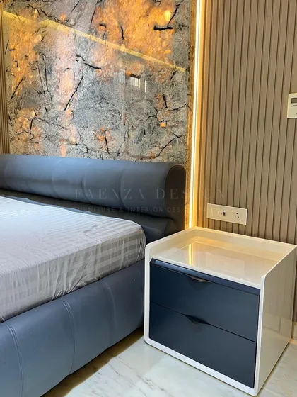 A close-up of a custom bedside table with a white top and dark blue drawers. It is paired with a leather-upholstered bed, set against a dramatic, back-lit stone-effect wall panel.