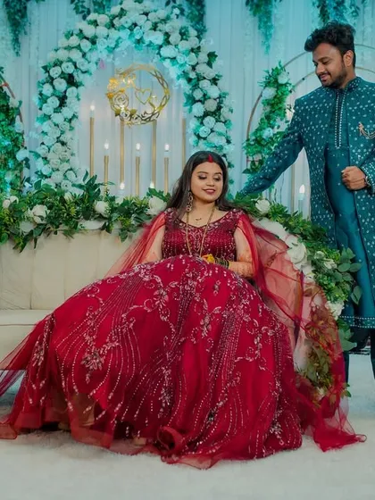 A stylish reception portrait. The groom stands beside his seated bride, whose elegant red gown flows around her, creating a grand and romantic image against the beautifully decorated stage.