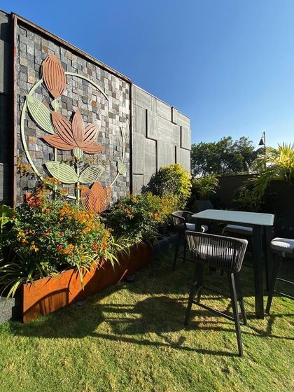 This art installation stands tall amidst a lush green garden, creating a beautiful backdrop for a high-top seating area. It enhances the aesthetic appeal of the landscape and provides a unique conversation piece.