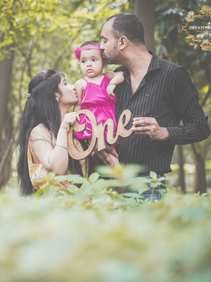 Parents kiss their one-year-old daughter, who is holding a "One" sign, in a lush green park.
