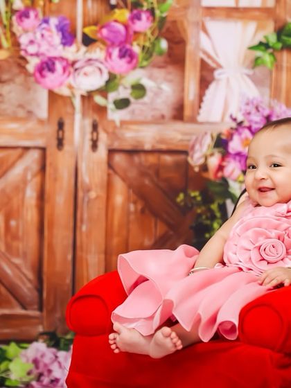 This little one is all smiles, sitting comfortably in a miniature red armchair. The floral backdrop adds a touch of rustic charm to her sitter portrait.