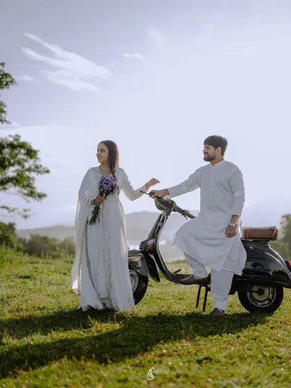 A retro-themed pre-wedding shoot featuring a classic black scooter. The all-white outfits and scenic mountain backdrop create a timeless, elegant look.