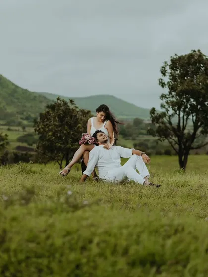 A romantic pre-wedding portrait with the groom lying in the grass and the bride sitting beside him in a serene, natural setting.