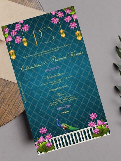 A vibrant and colorful traditional wedding invitation. This design uses a rich teal background with a classic Indian pattern, brought to life with bright pink lotuses, hanging lanterns, and a majestic peacock.