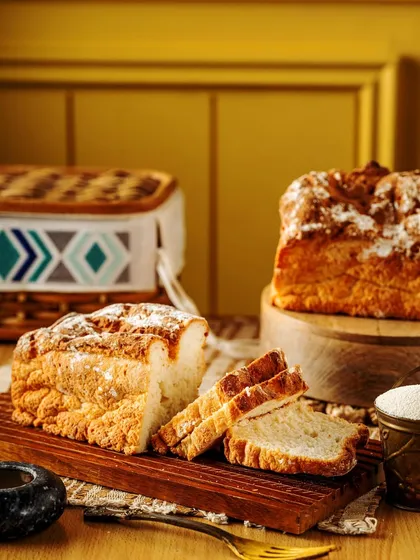 A rustic loaf of artisan bread, sliced to show its soft interior. The warm, home-style setup with a wooden board and picnic basket evokes a feeling of comfort and traditional baking.