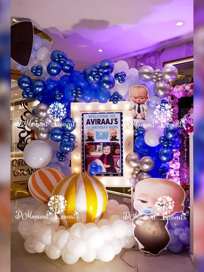 A fun and inviting entrance for a Boss Baby themed party. The setup includes a lighted welcome sign, character cutouts, and a balloon arrangement in blue, white, and silver, with striped hot air balloon props.
