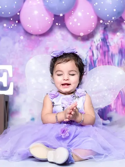 Clapping her hands with joy in her fairy costume. The combination of the purple dress, delicate wings, and happy expression makes this a truly magical first birthday photo.