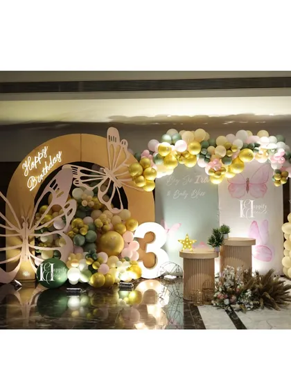 A whimsical butterfly-themed birthday party setup for a third birthday. The design includes large, custom-cut butterfly wings, a "Happy Birthday" neon sign, and an organic balloon arrangement in pastel green, gold, and pink, creating a magical photo corner.